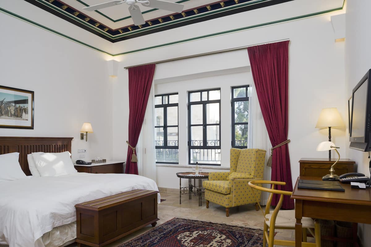 A hotel room with a bed, a desk with a lamp, a chair, a small table, and a window with curtains.