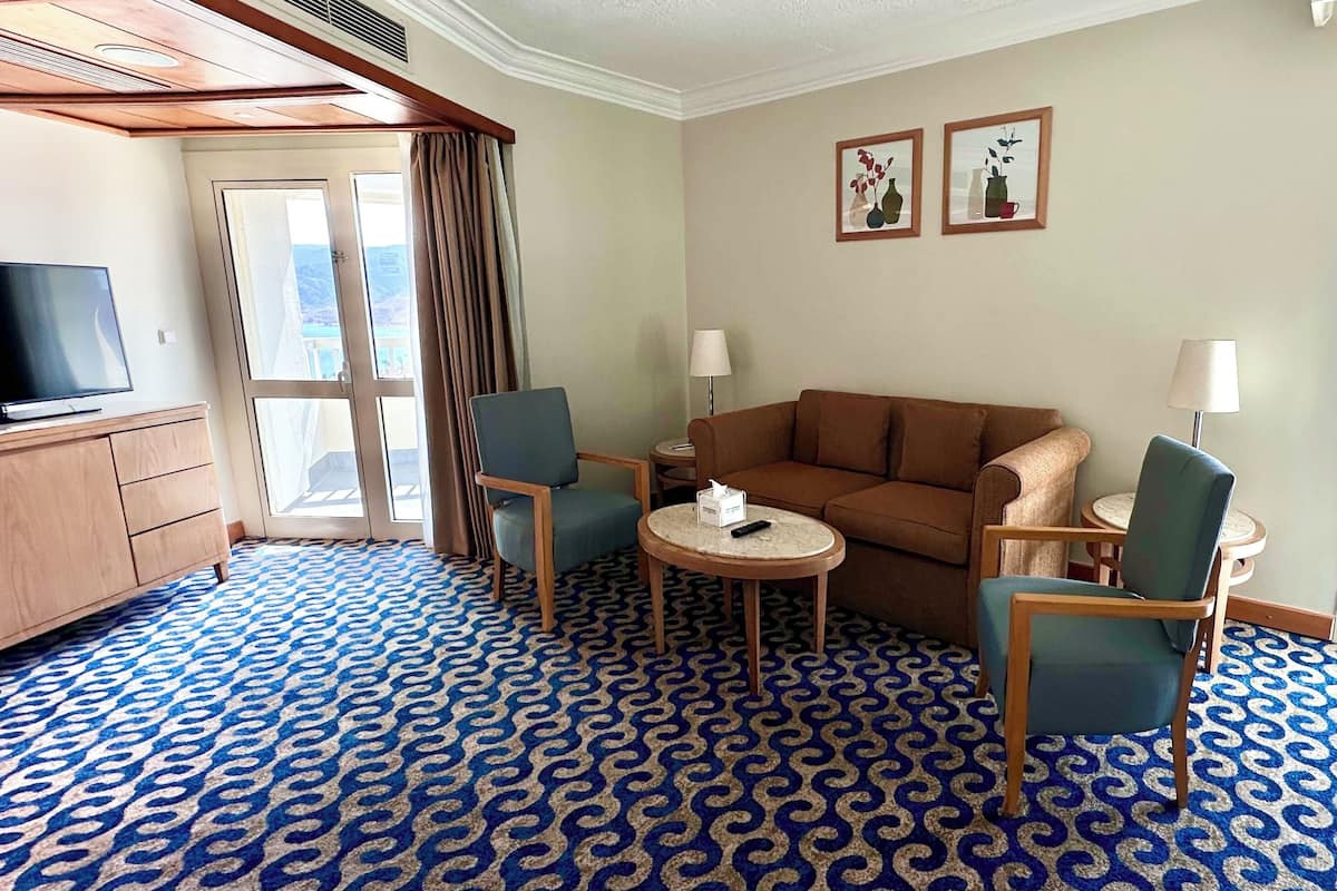 Hotellihuone, jossa on suuri televisio, sohva, kaksi nojatuolia, pieni pöytä ja suuri ikkuna, josta avautuu näkymä vuorille.