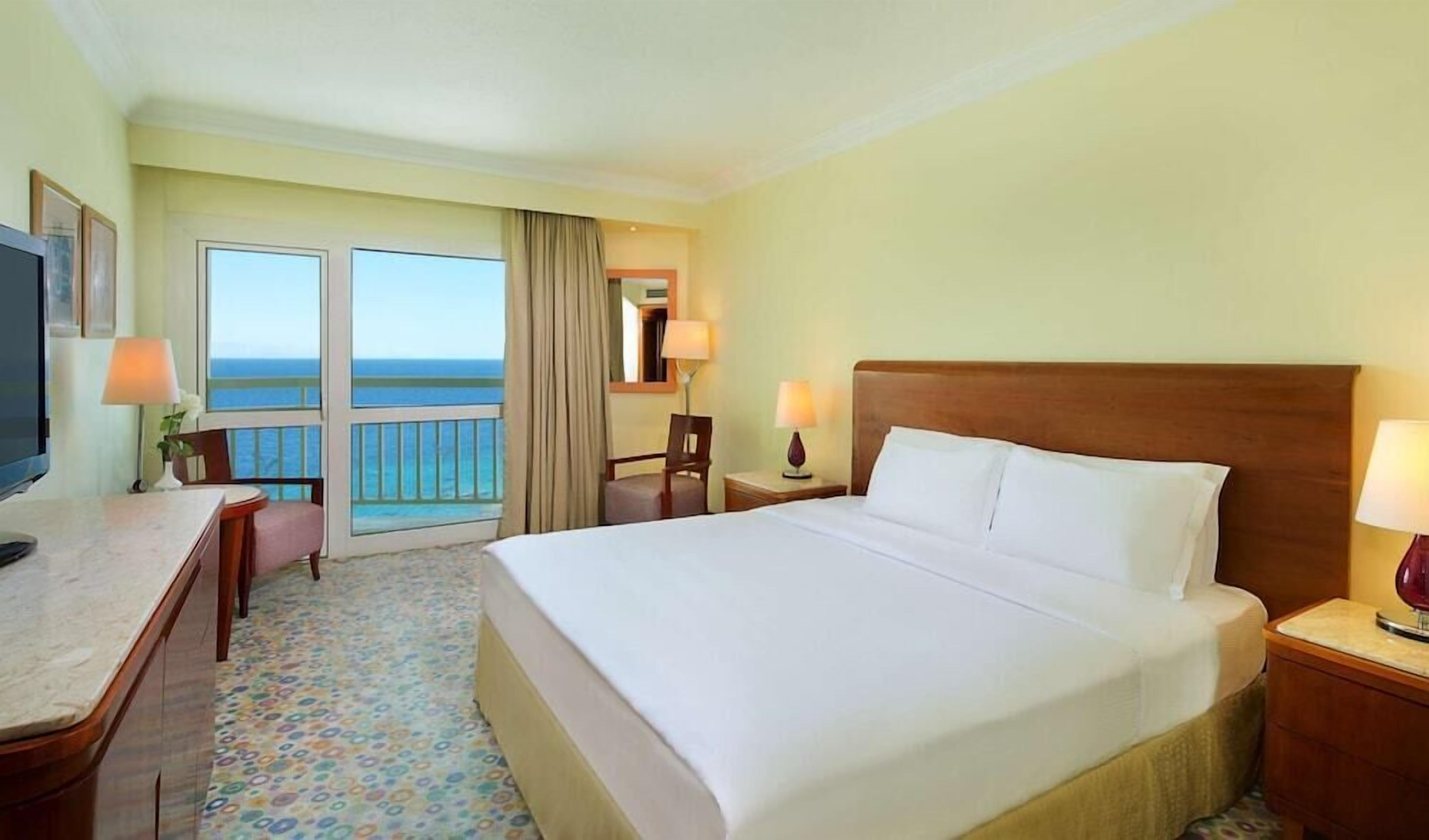 A hotel room with a bed, a desk, a chair, a TV, and a view of the ocean.