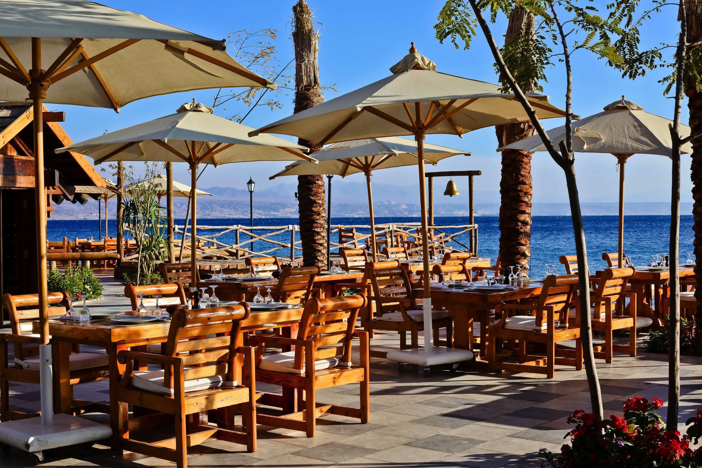 Un área de comedor al aire libre con mesas y sillas de madera, sombrillas color beige y vista al mar.