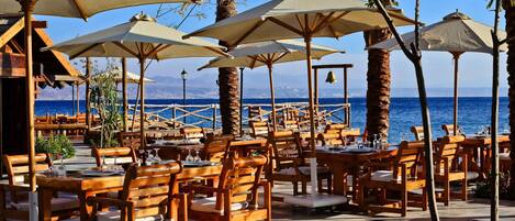 Un área de comedor al aire libre con mesas y sillas de madera, sombrillas color beige y vista al mar.