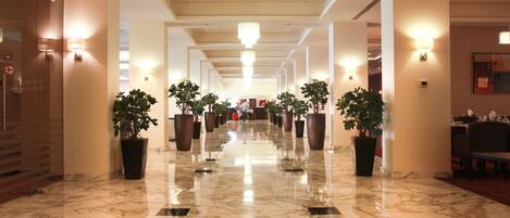 Un pasillo de hotel con pisos de mármol, plantas en macetas, candelabros y apliques.