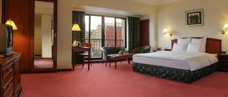 Habitación de hotel con una cama grande, un escritorio con silla, una pequeña mesa con fruta, televisión y balcón con vistas a edificios.
