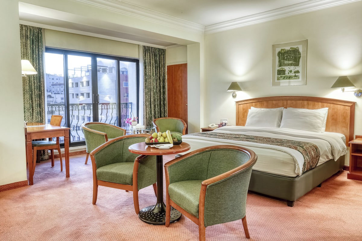 A hotel room with a bed, a dining table with chairs, a small table with fruit, and a balcony with curtains.