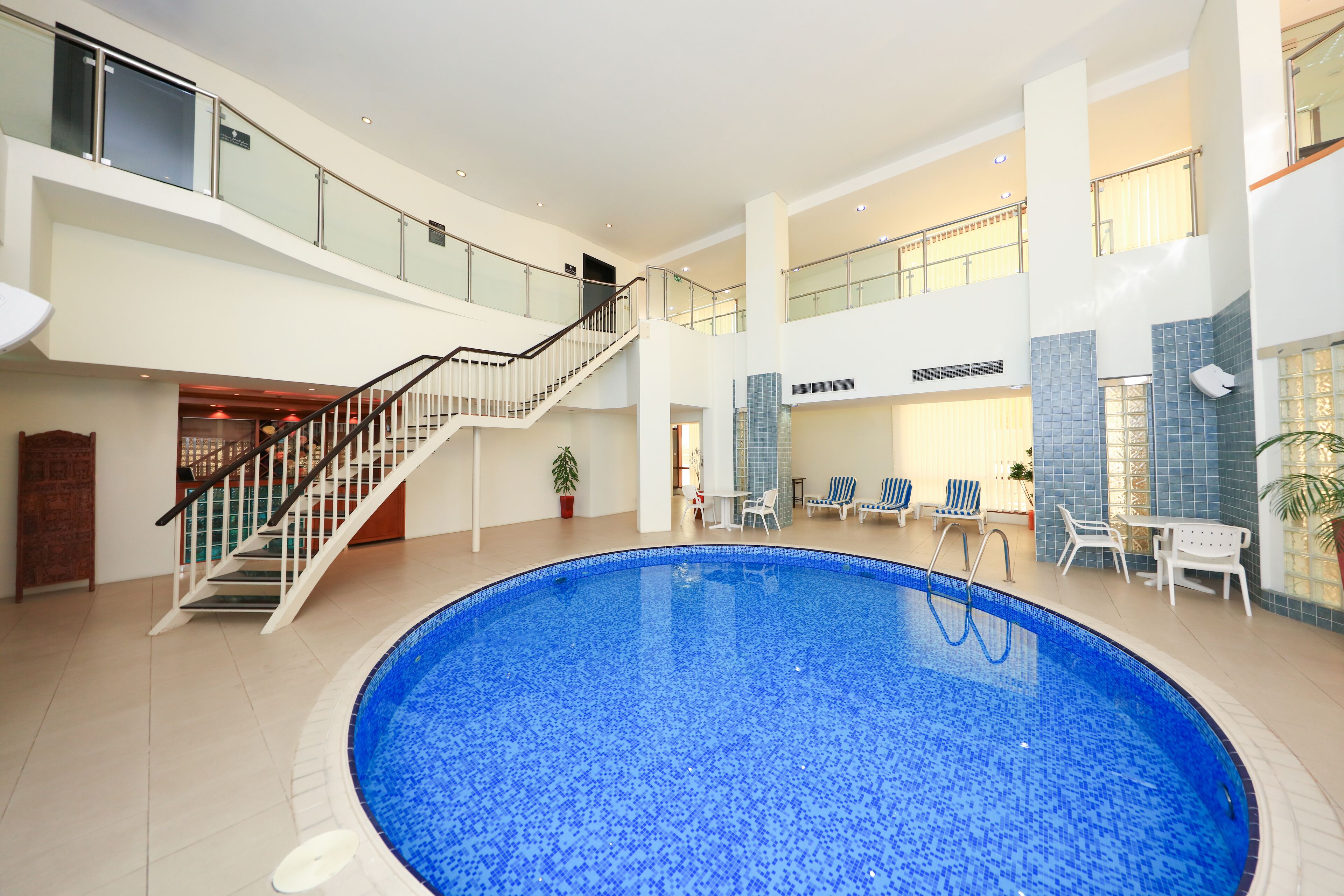Una amplia piscina interior con balaustrada de vidrio, una escalera y sillones de descanso.