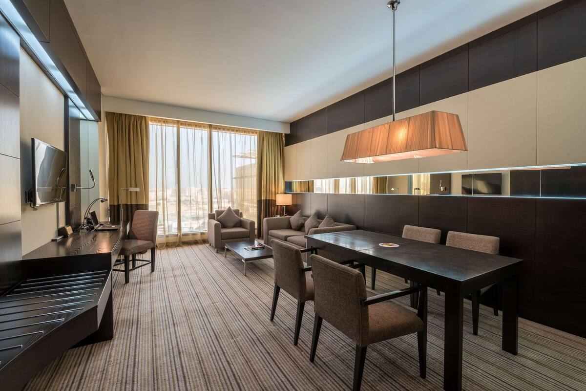 A modern hotel room with a piano, a dining table with chairs, a sofa, and a large window with curtains.