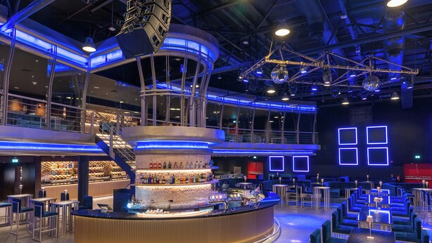 A modern bar area with a central bar counter, seating arrangements, and a staircase leading to an upper level.