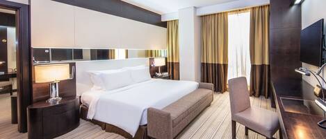 A modern hotel room with a large bed, a desk with a chair, a flat-screen TV, and a large mirror.