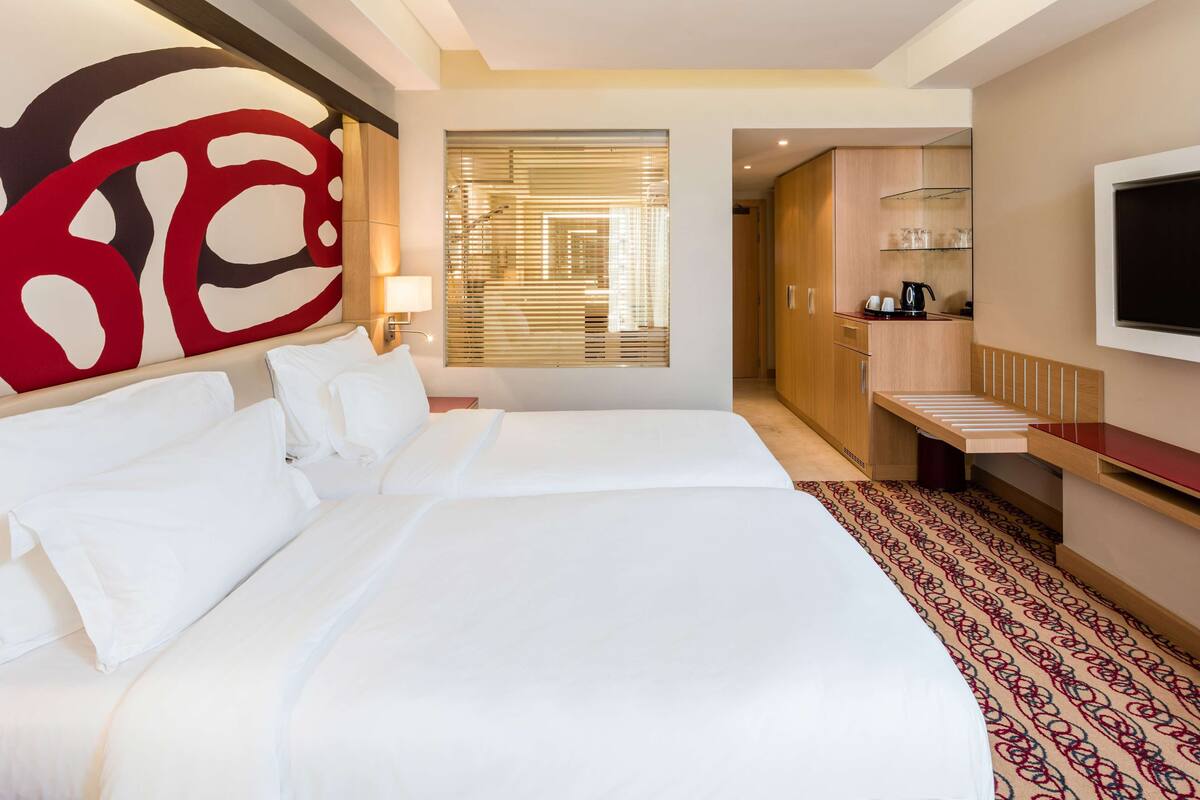 A hotel room with two beds, a large abstract wall art piece, a TV mounted on the wall, and a wooden wardrobe.