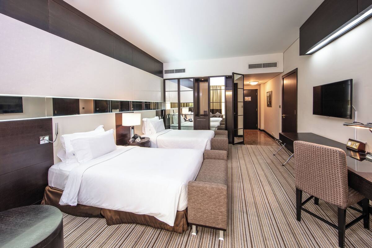A hotel room with two beds, a desk, a chair, a TV, and a bathroom visible through an open door.