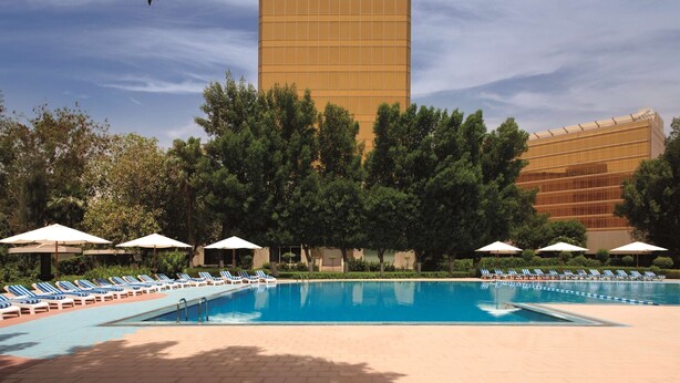 A swimming pool with lounge chairs and umbrellas, surrounded by trees and a tall building in the background.