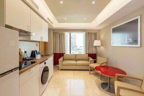A modern hotel room with a kitchenette, a sofa, a dining table, and a large window with curtains.