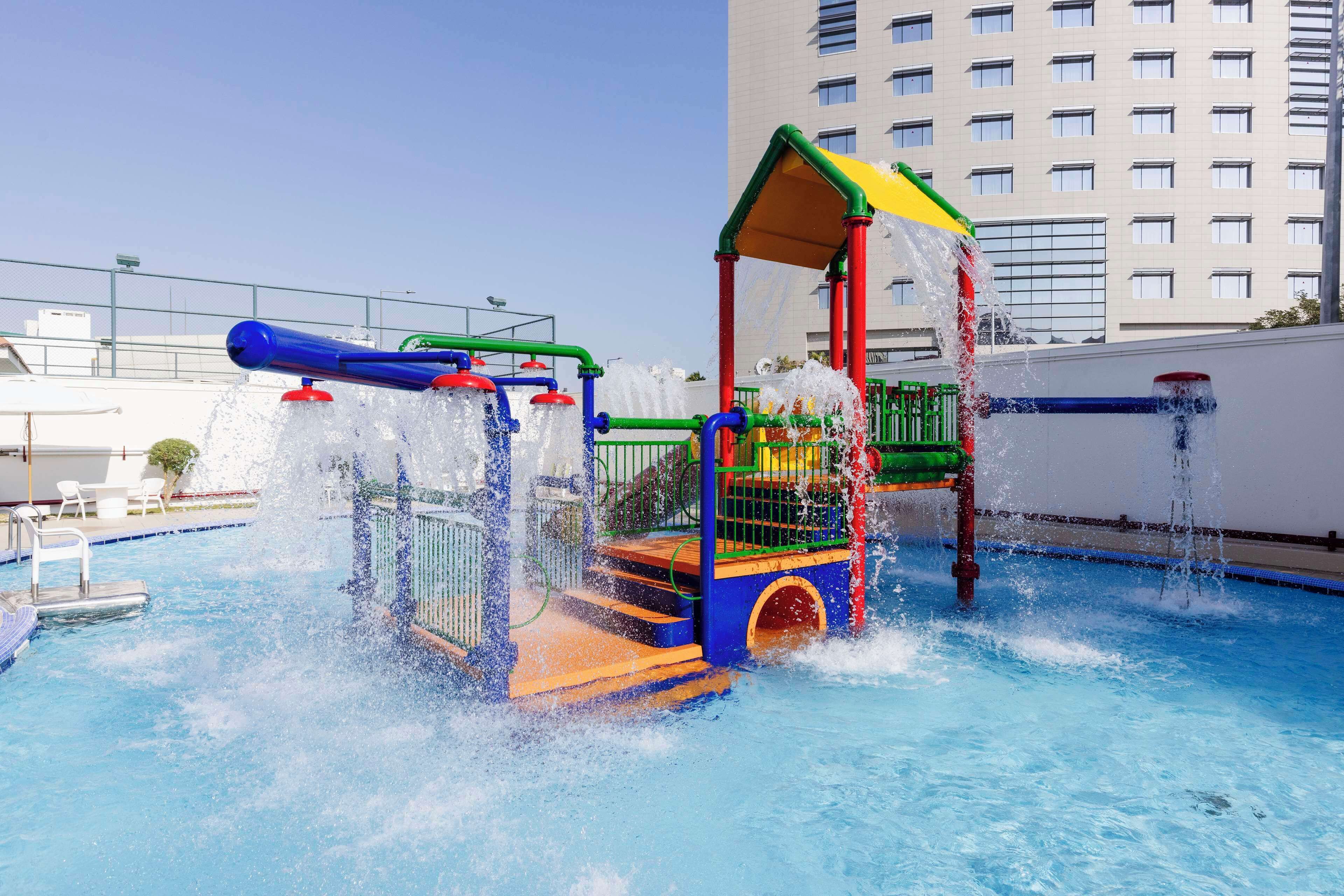 A water play area with a slide and splash pad, surrounded by a pool and a high-rise building in the background.