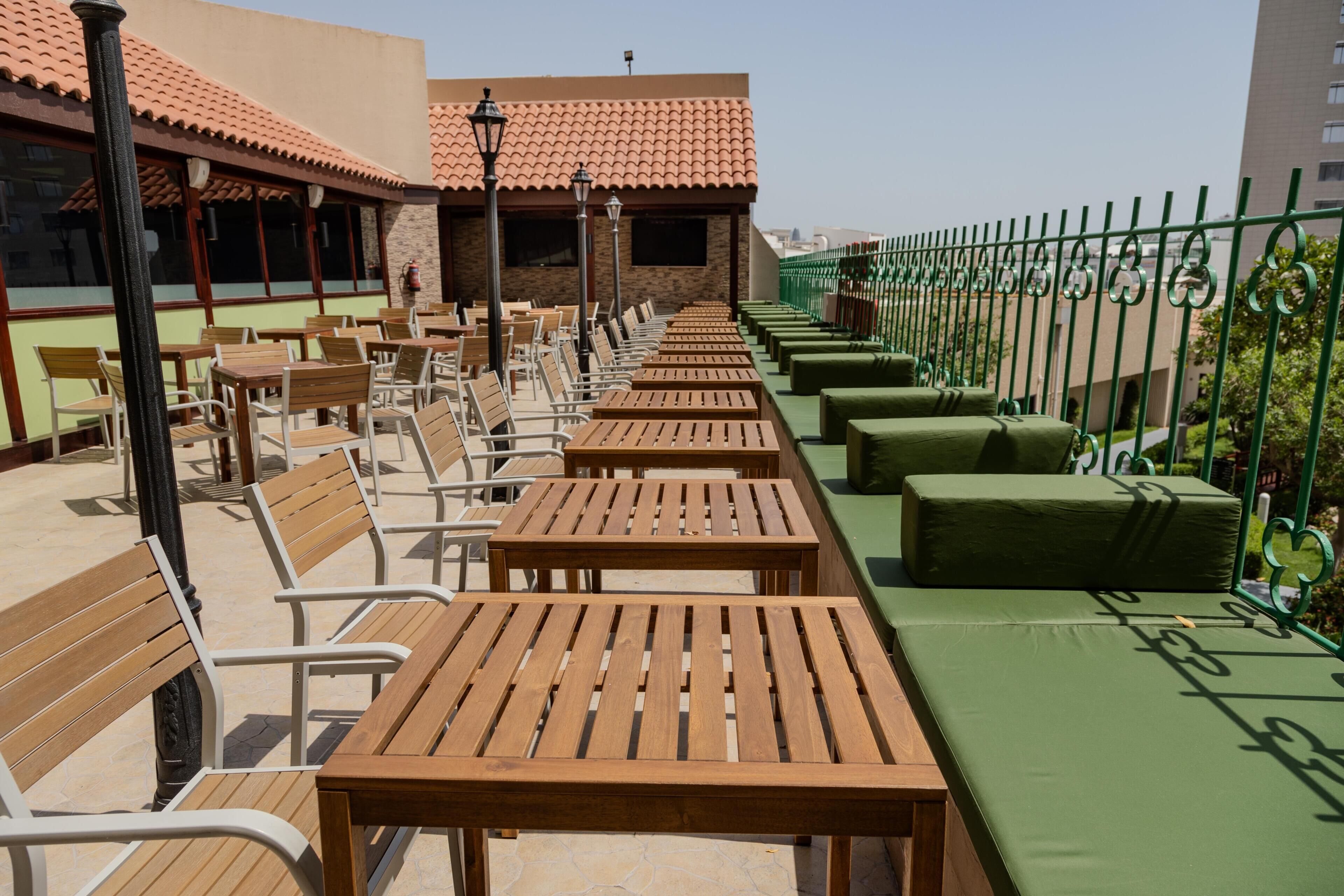 Una terraza con mesas y sillas de madera, un área de estar con cojines verdes y un techo de tejas.