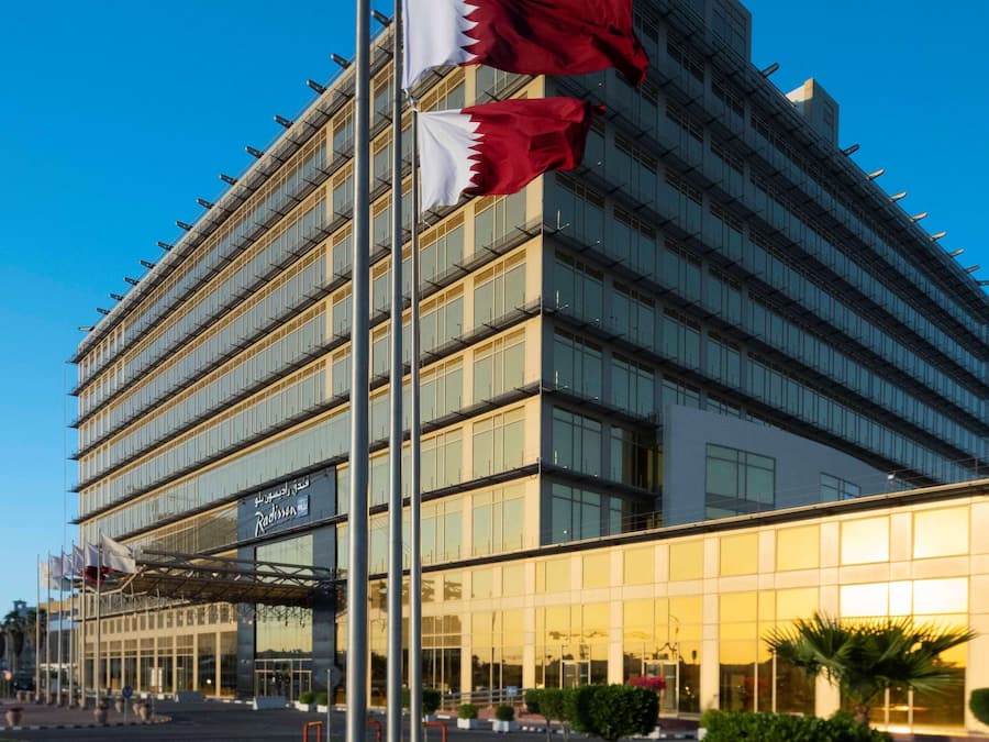 A modern hotel with multiple flags on a flagpole, large glass windows, and a well-maintained lawn.