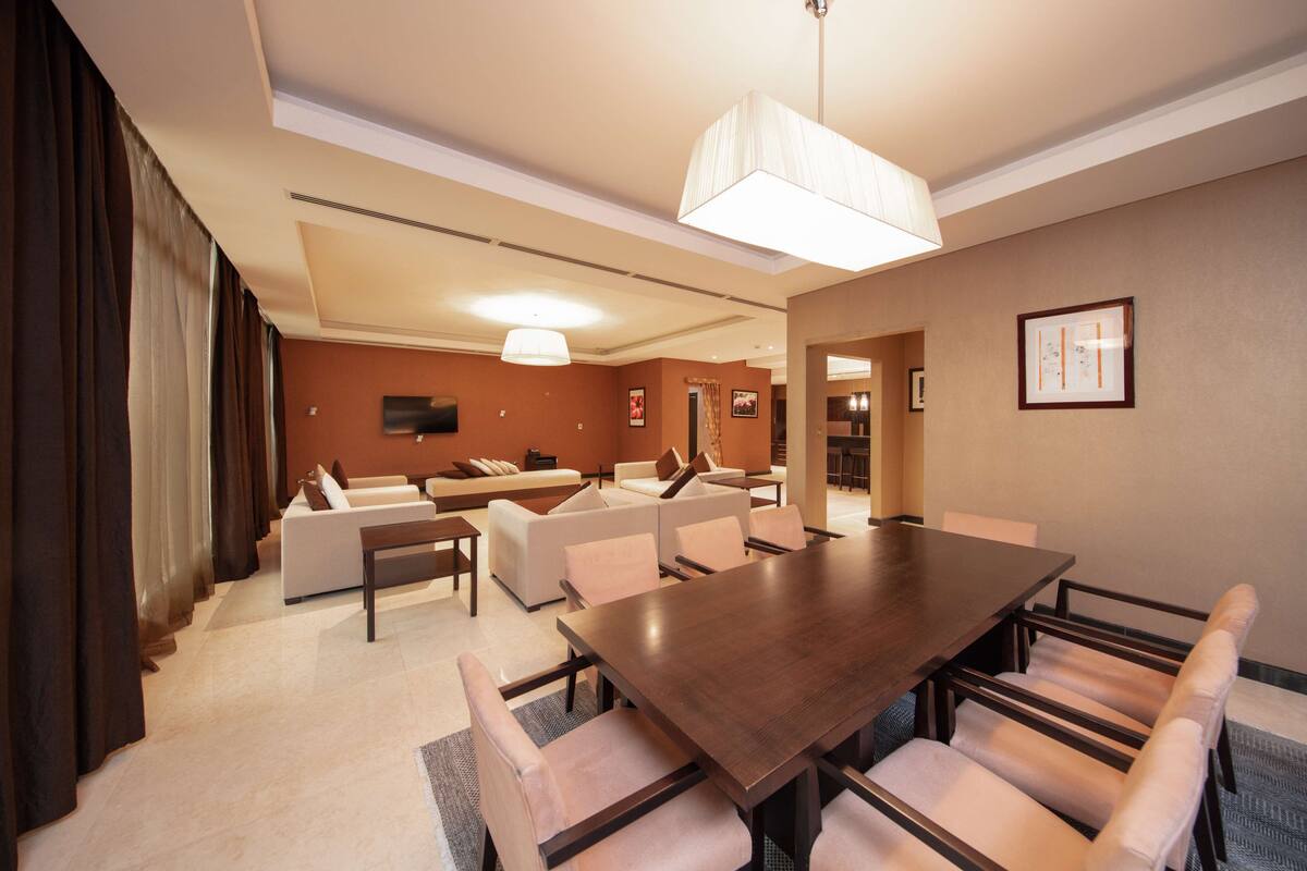 A modern hotel room with a large dining table, multiple sofas, and a large window with curtains.