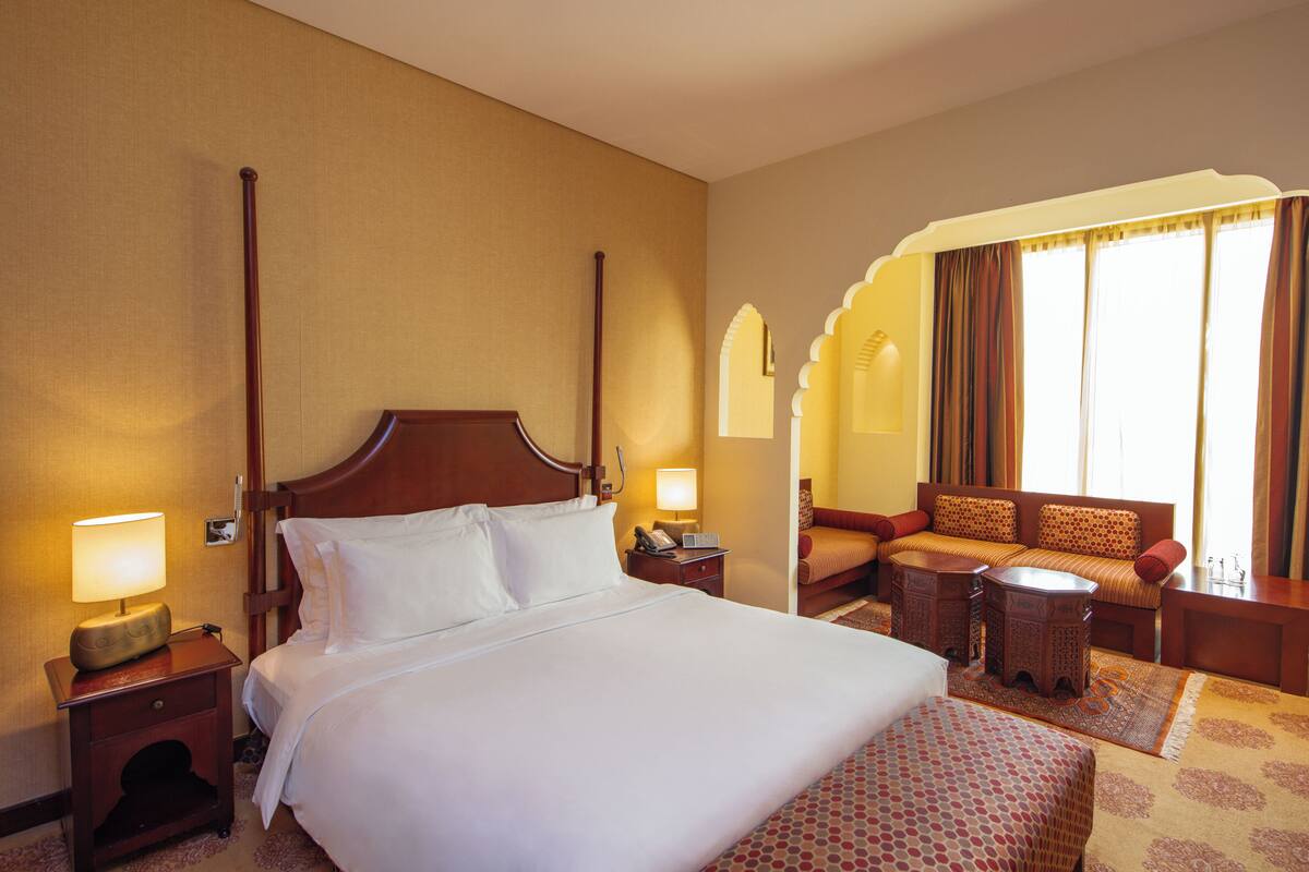 A hotel room with a large bed, a sofa, a small table, and a nightstand with a lamp.