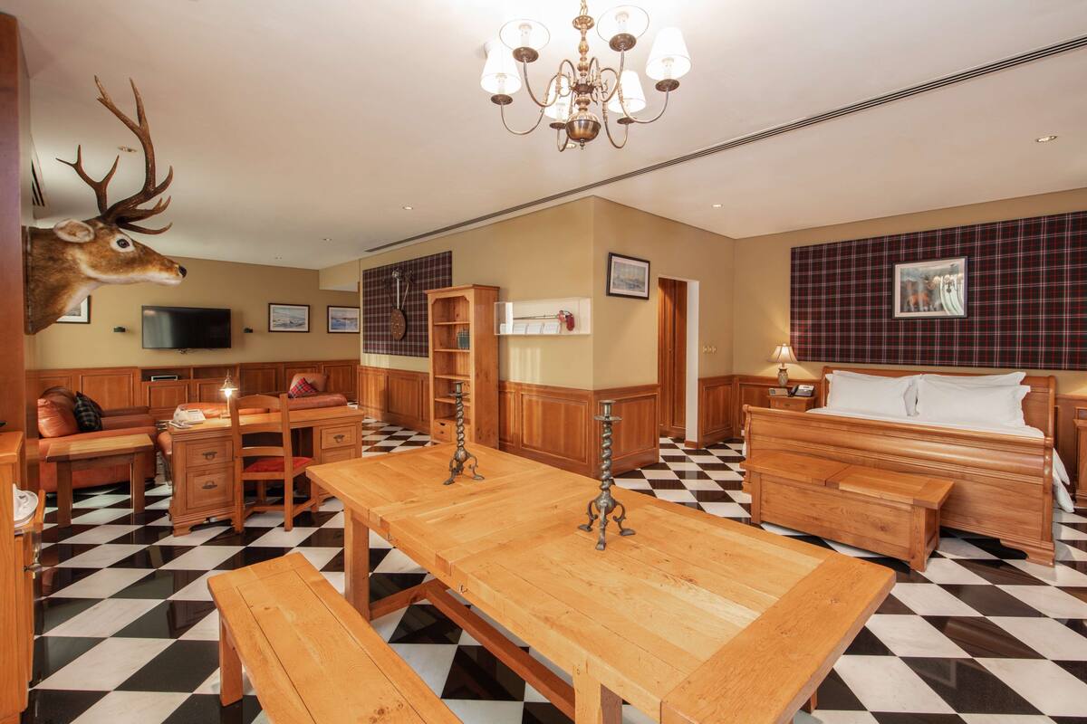 A hotel room with a checkered floor, wooden furniture, a mounted deer head, a chandelier, and a bed with a plaid headboard.