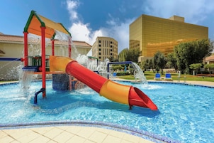 Parque acuático con un tobogán rojo y amarillo, una piscina y un edificio moderno al fondo.