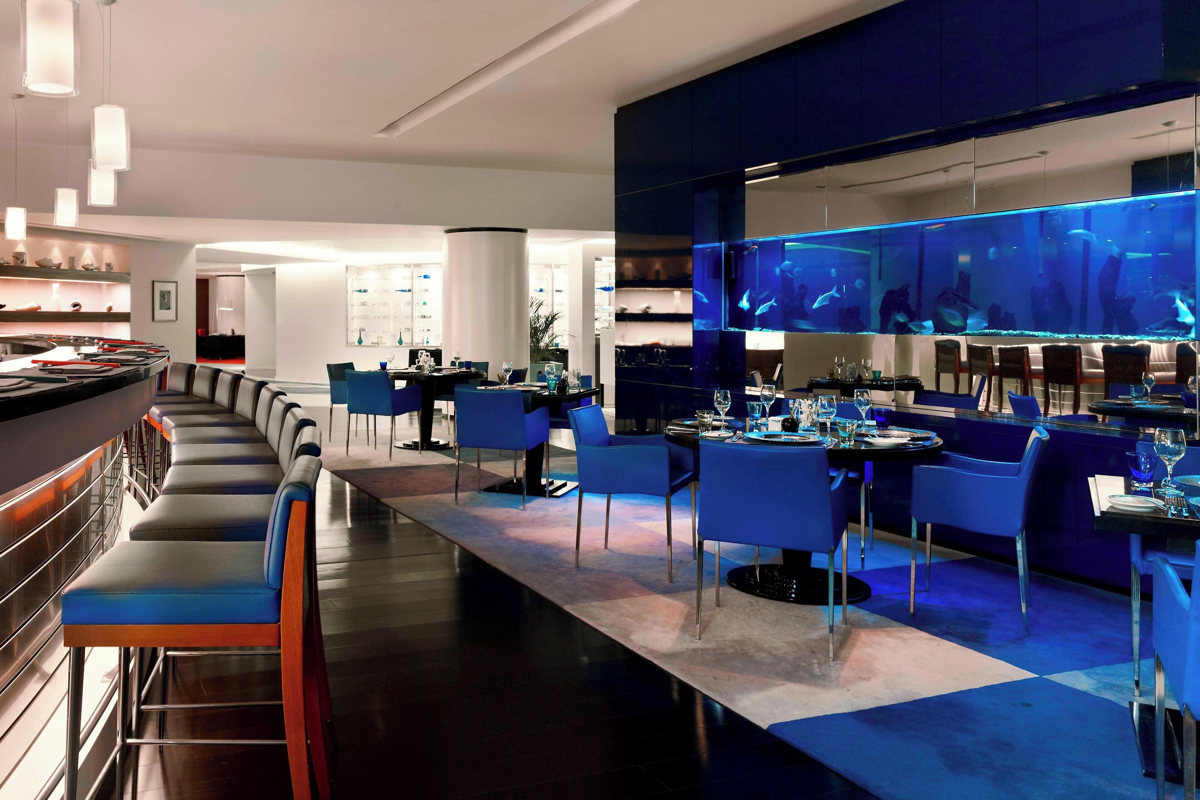 A modern restaurant with a long bar, blue chairs, and a large aquarium.