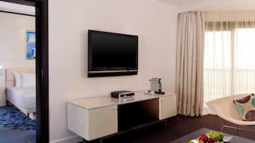 A modern hotel room with a bed, a TV mounted on the wall, a coffee machine, and a dining table with fruit.