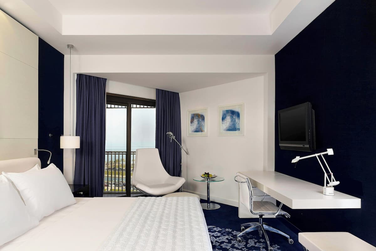 A hotel room with a bed, a desk, a chair, a TV, and a balcony with curtains.