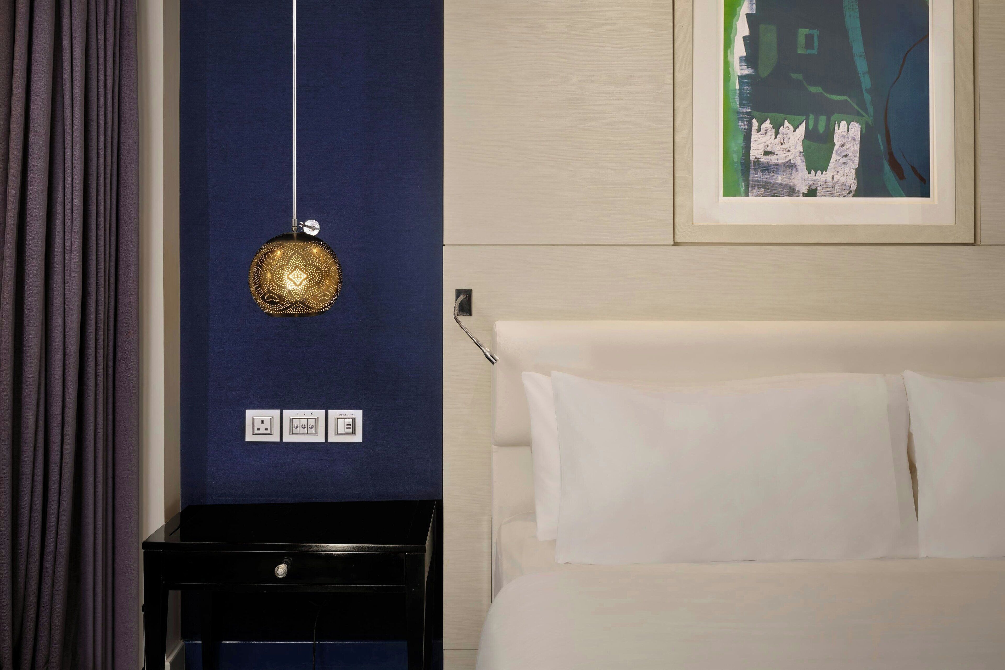 A hotel room with a bed, a nightstand, a wall-mounted light fixture, and a framed picture on the wall.