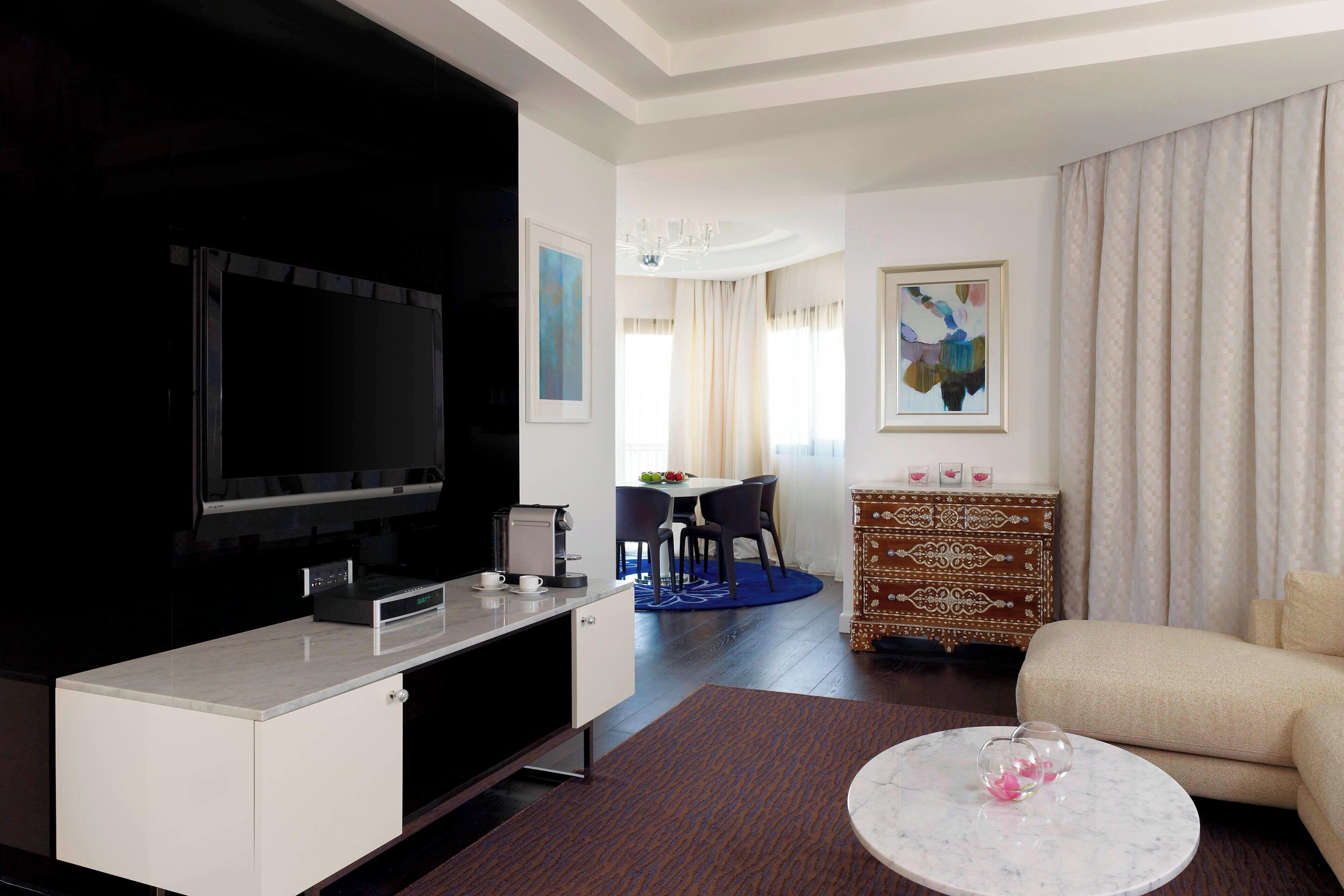 Una habitación de hotel moderna con televisor de pantalla plana, un área de comedor con mesa y sillas, un sofá, un aparador estampado y una araña.