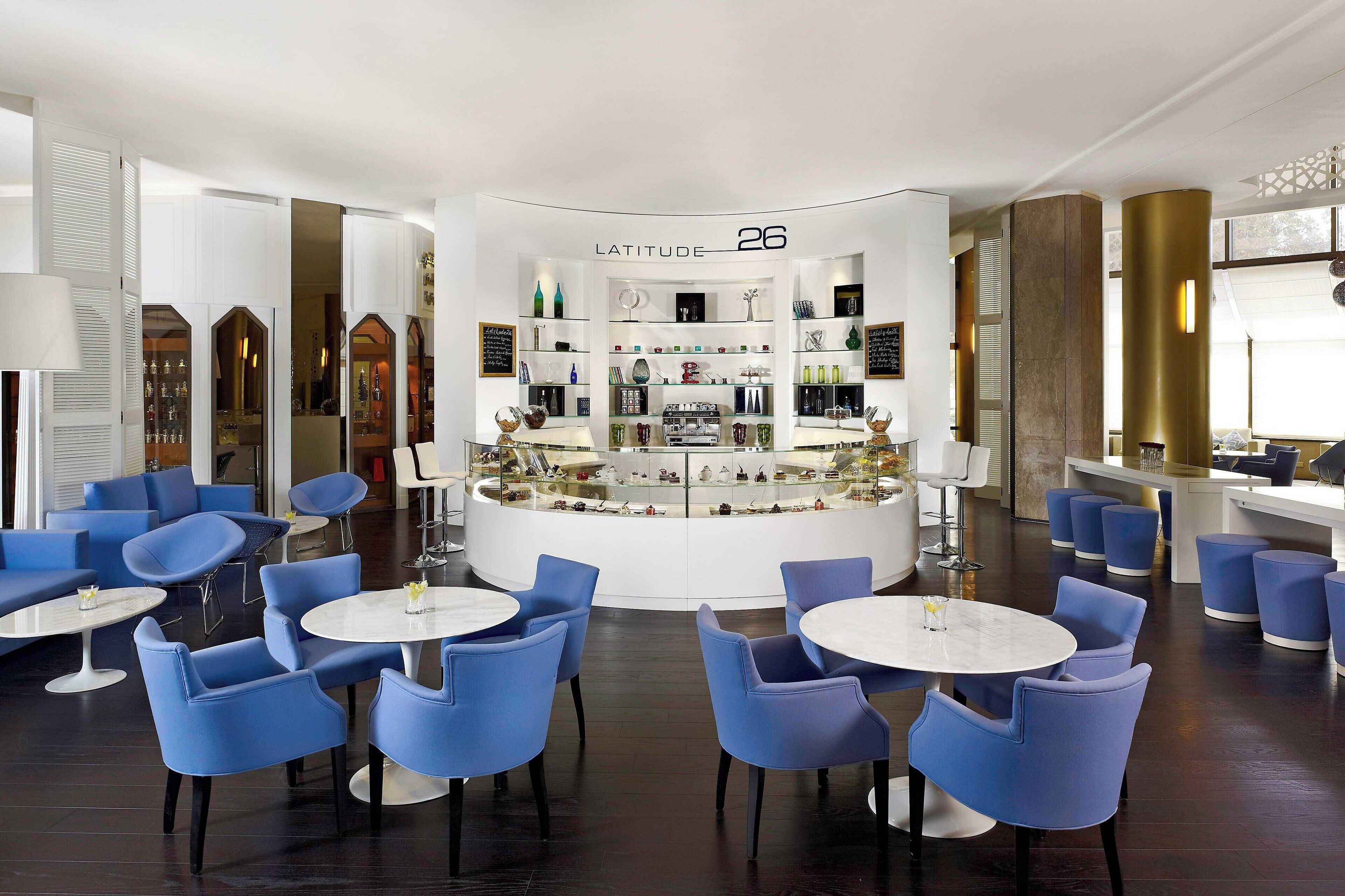 A modern lounge area with blue chairs and tables, a central display case labeled "LATITUDE 26", and a bar area with stools.