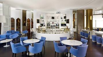 A modern lounge area with blue chairs and tables, a central display case labeled "LATITUDE 26", and a bar area with stools.