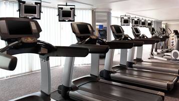 Varias caminadoras en un gimnasio con ventanas amplias y múltiples pantallas colgantes.