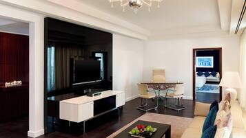 A modern living room with a flat-screen TV, a coffee table with fruit, a sofa with cushions, a dining area with a table and chairs, and a hallway leading to a bedroom.