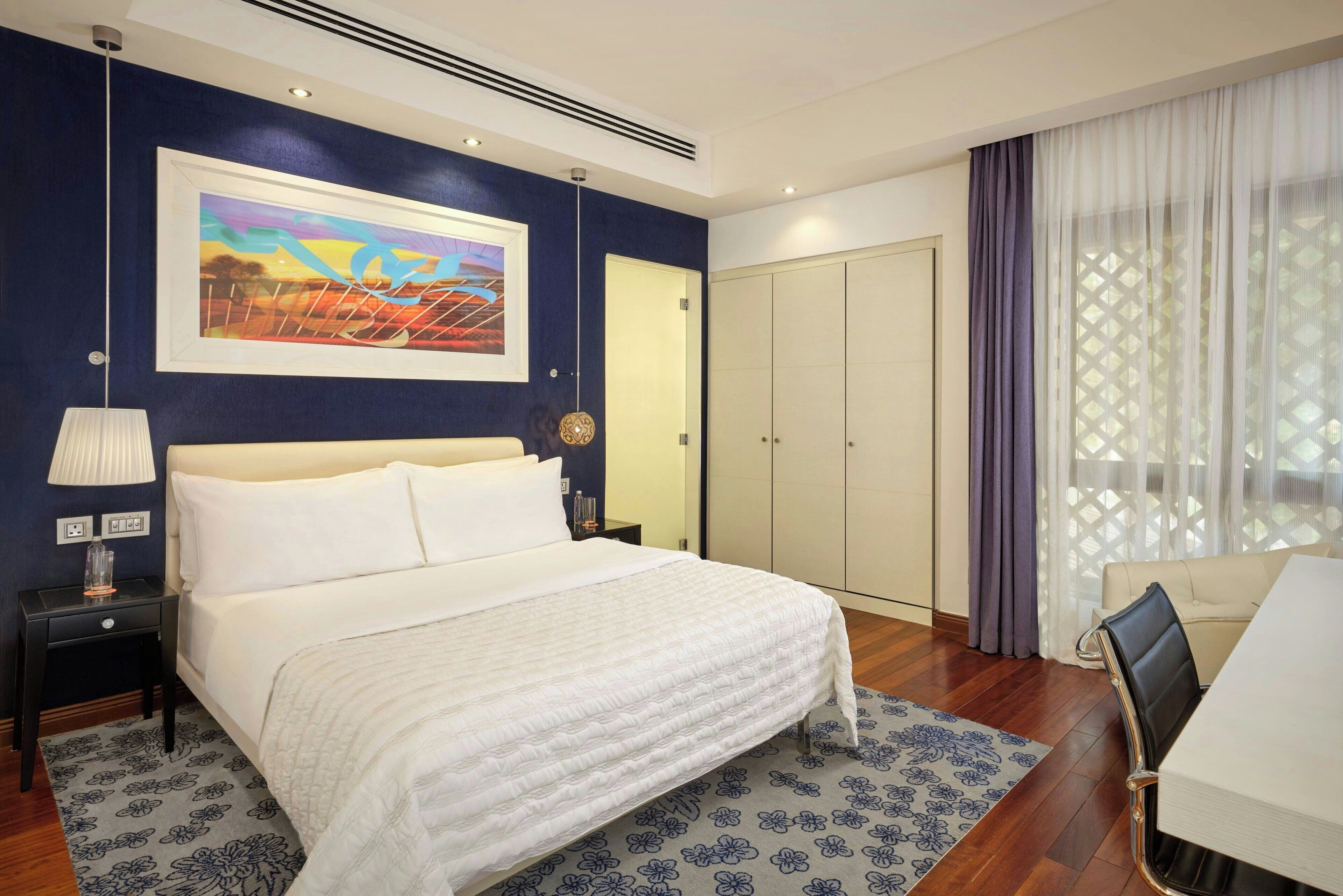 Une chambre d’hôtel avec un grand lit, une table de chevet avec une lampe, un tableau au mur, une armoire et un coin repas avec une table et des chaises.