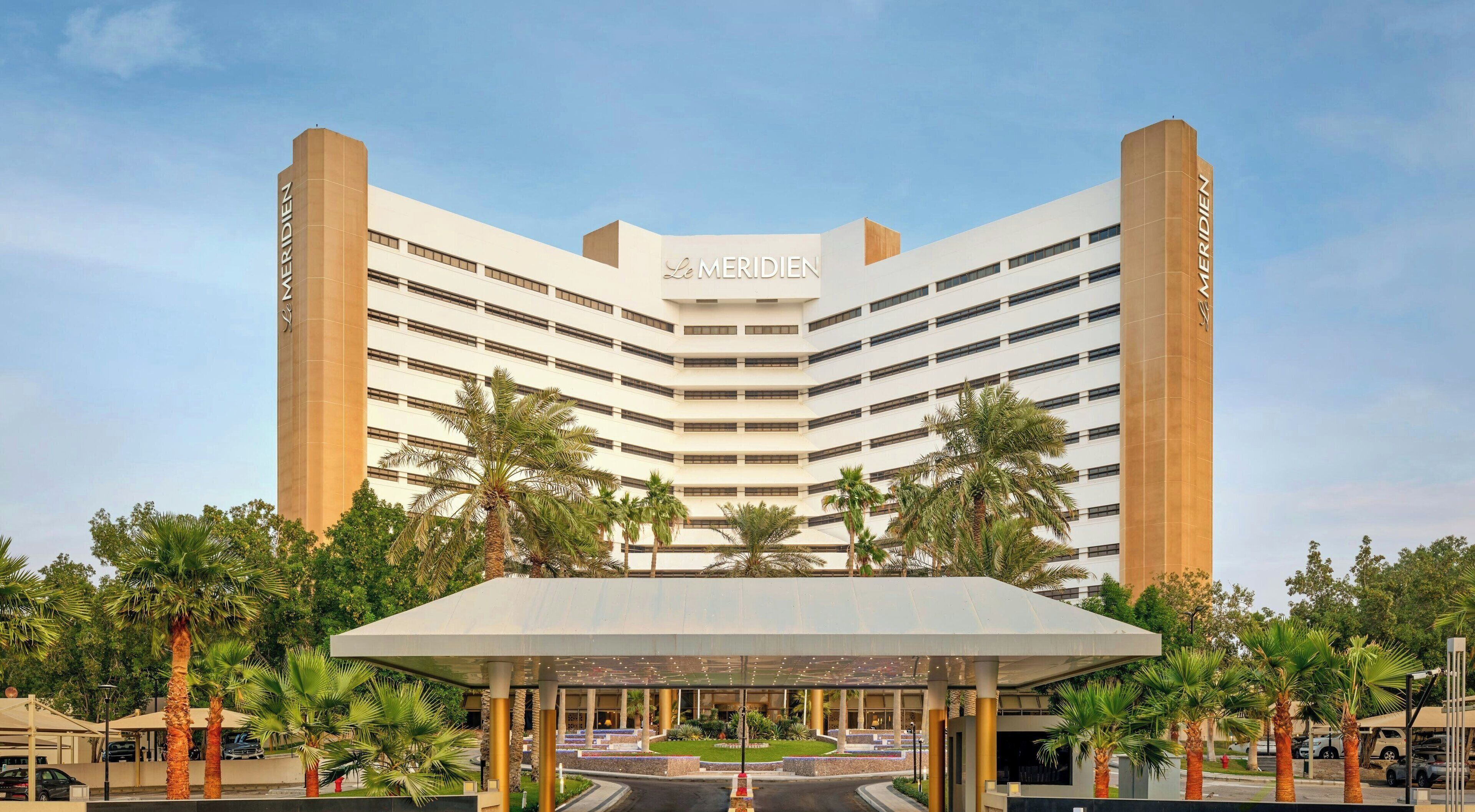Un hotel moderno con un llamativo letrero de 'Le Meridien', que presenta una estructura de varios pisos con un atrio central y palmeras en primer plano.