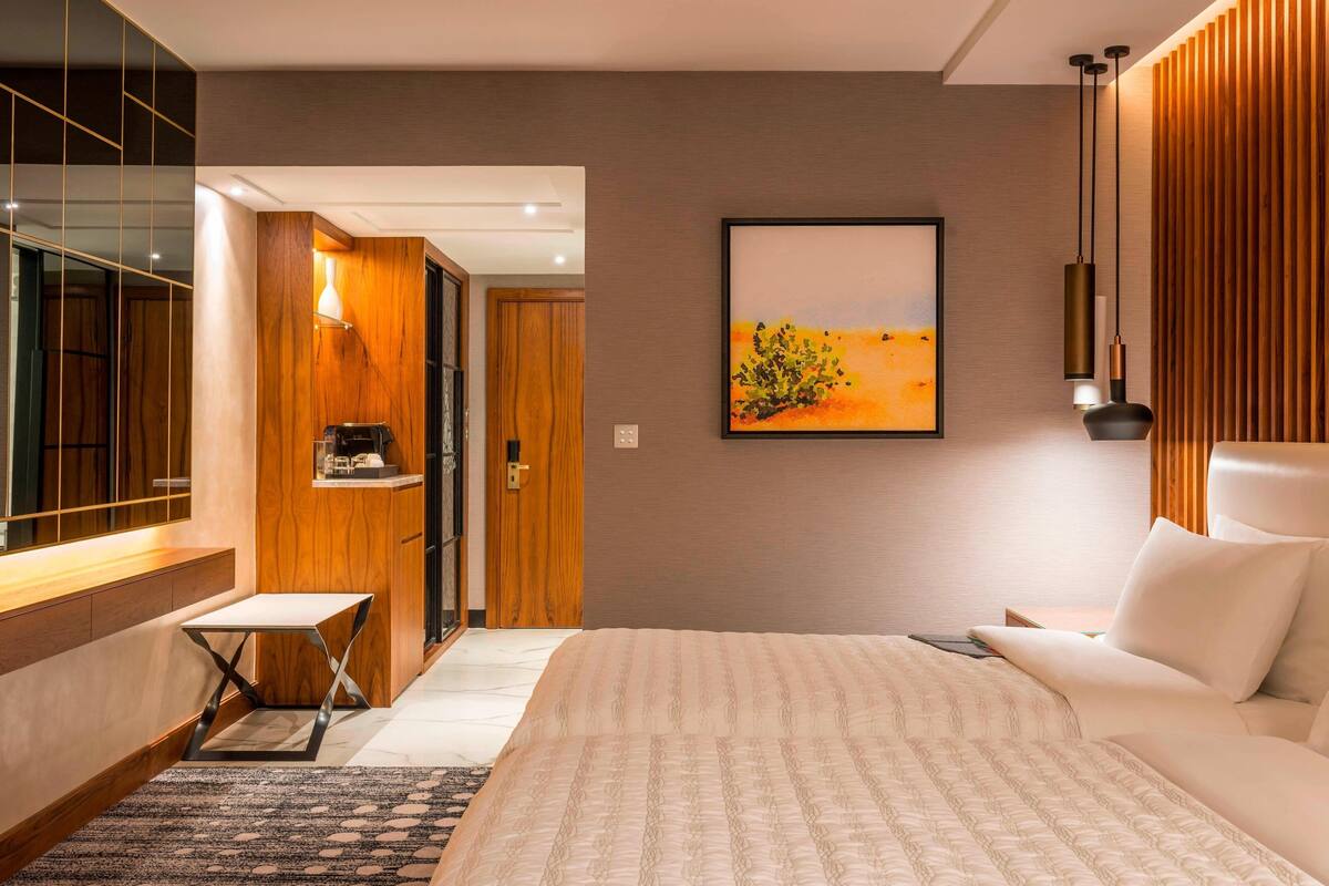 A modern hotel room with a large bed, a small table, a desk, and a painting on the wall.