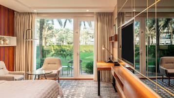 Habitación de hotel con cama, dos sillones, una mesita, televisor y un ventanal que ofrece vistas a palmeras y un jardín.