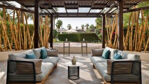 A covered outdoor seating area with two sofas and a central table, surrounded by bamboo pillars and a view of palm trees and buildings. - Le Meridien Dubai Hotel & Conference Centre (Dubai)