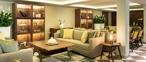 A modern living room with a sofa, coffee table, and books. There is a wooden cabinet with glass doors and a potted plant.