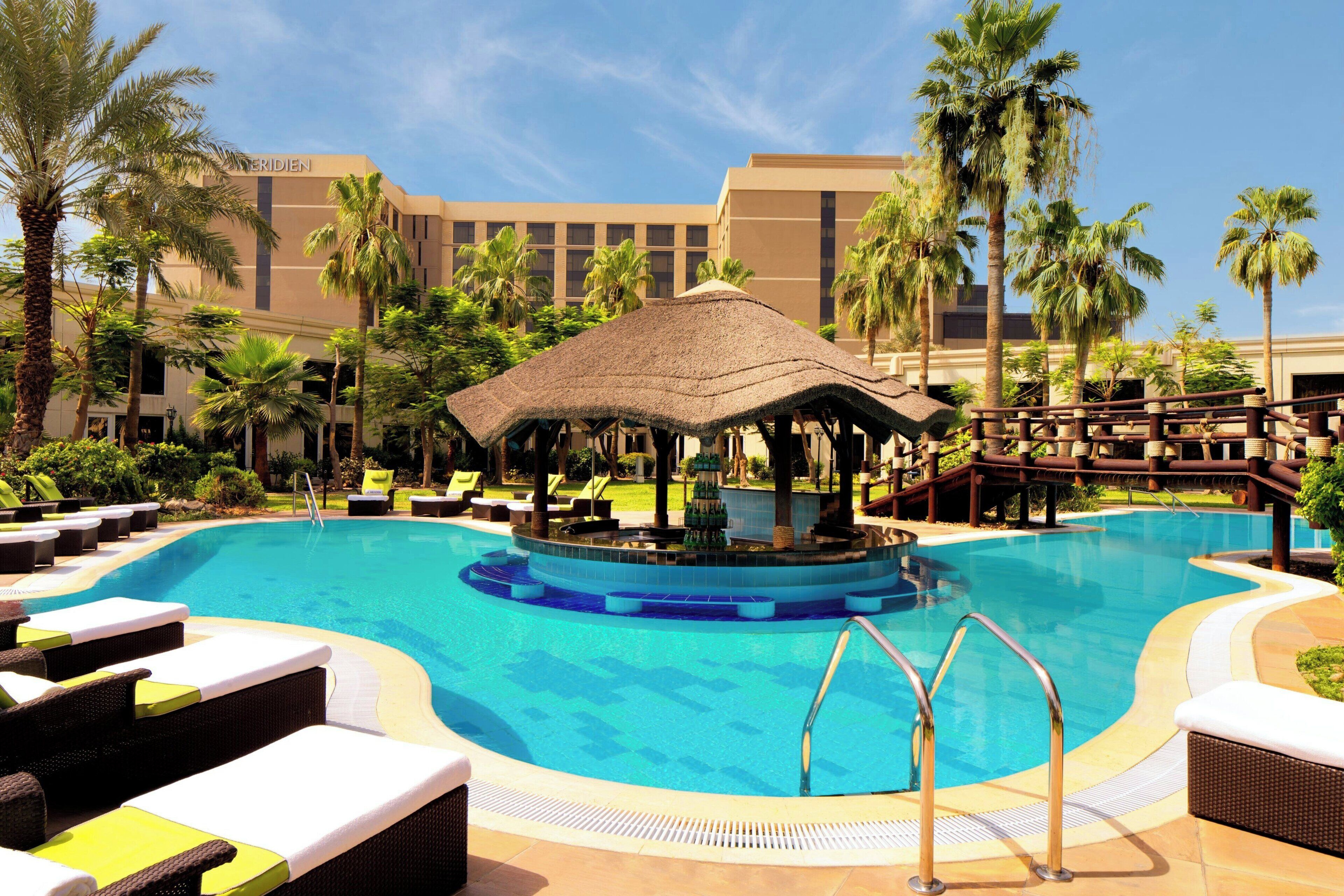 A resort pool area with lounge chairs, a central water feature, and palm trees.
