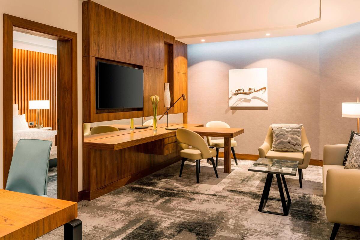 A hotel room with a wooden accent wall, a flat-screen TV mounted above a wooden desk, a dining area with a table and chairs, and a seating area with two armchairs and a sofa.