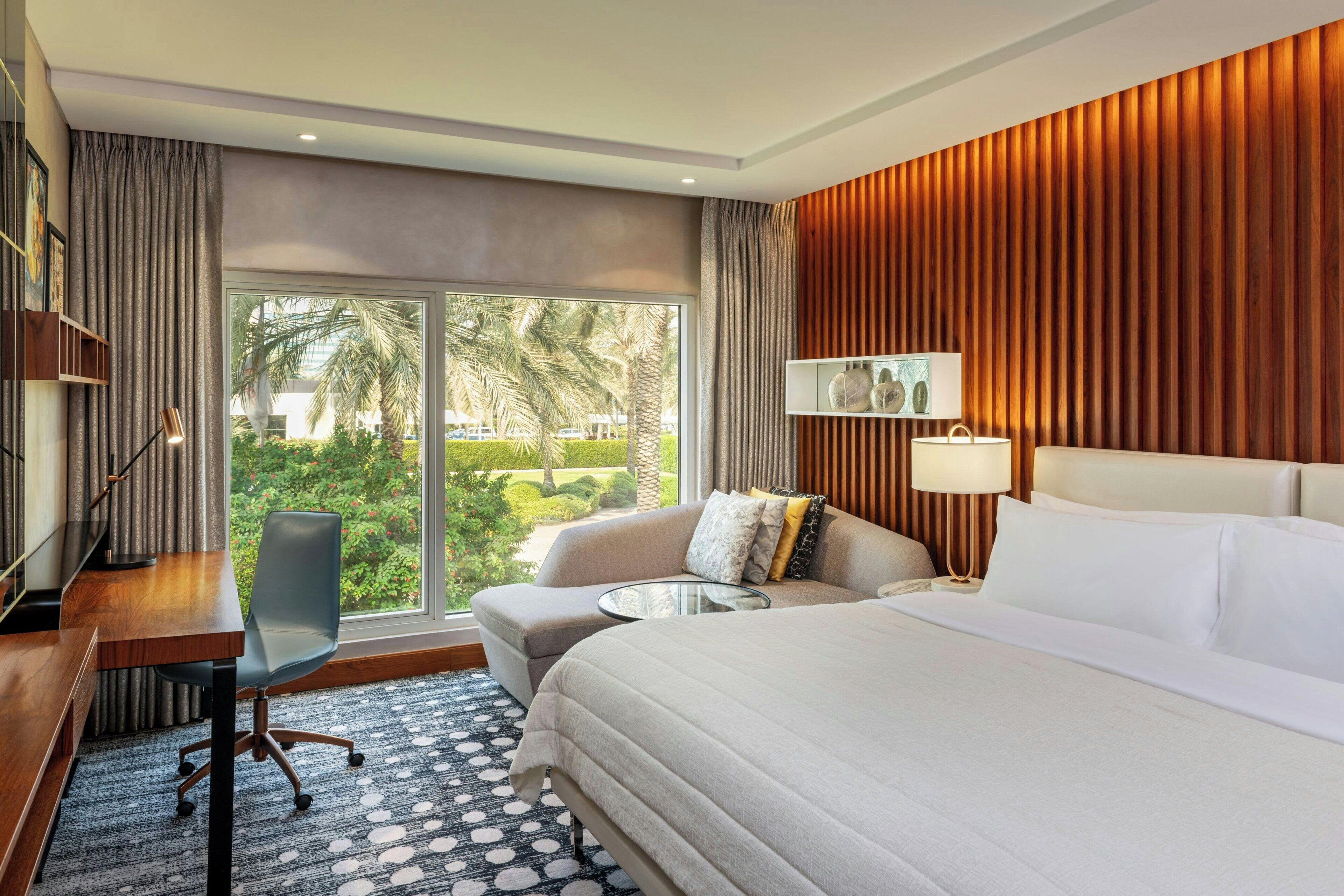 A hotel room with a large bed, a sofa, a desk, and a chair. There is a large window with a view of palm trees and a garden.