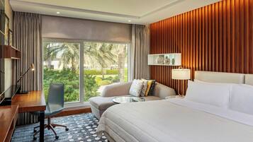 A hotel room with a large bed, a sofa, a desk, and a chair. There is a large window with a view of palm trees and a garden.