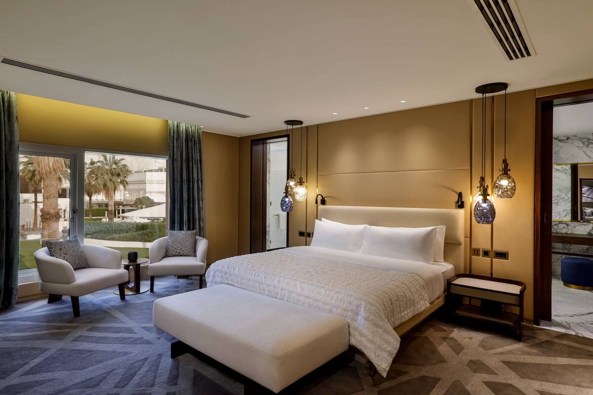 A modern hotel room with a large bed, two armchairs, a small table, and a bench.