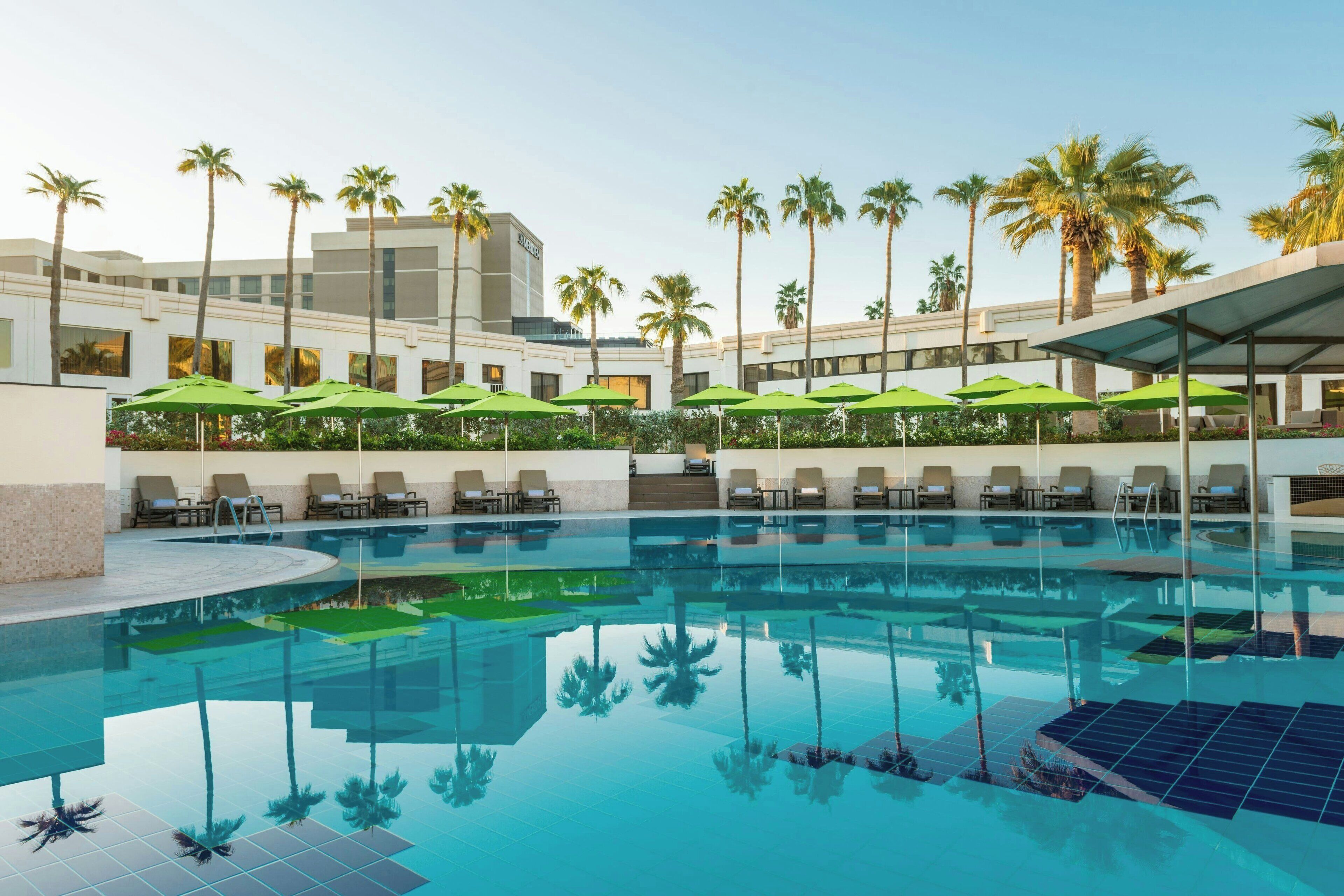 Ein Swimmingpool mit Liegestühlen und Sonnenschirmen, umgeben von Palmen und modernen Gebäuden.
