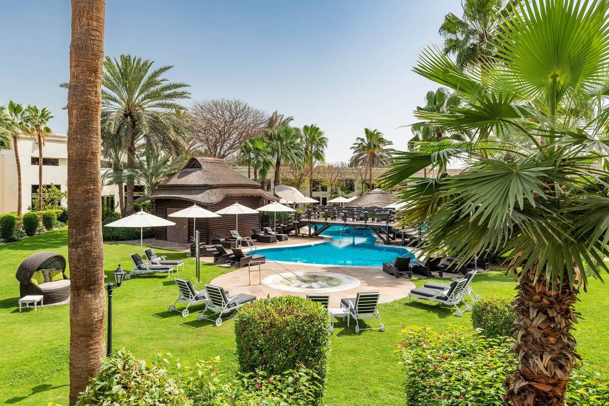 A pool area with lounge chairs and umbrellas, surrounded by palm trees and a building with a dome roof.
