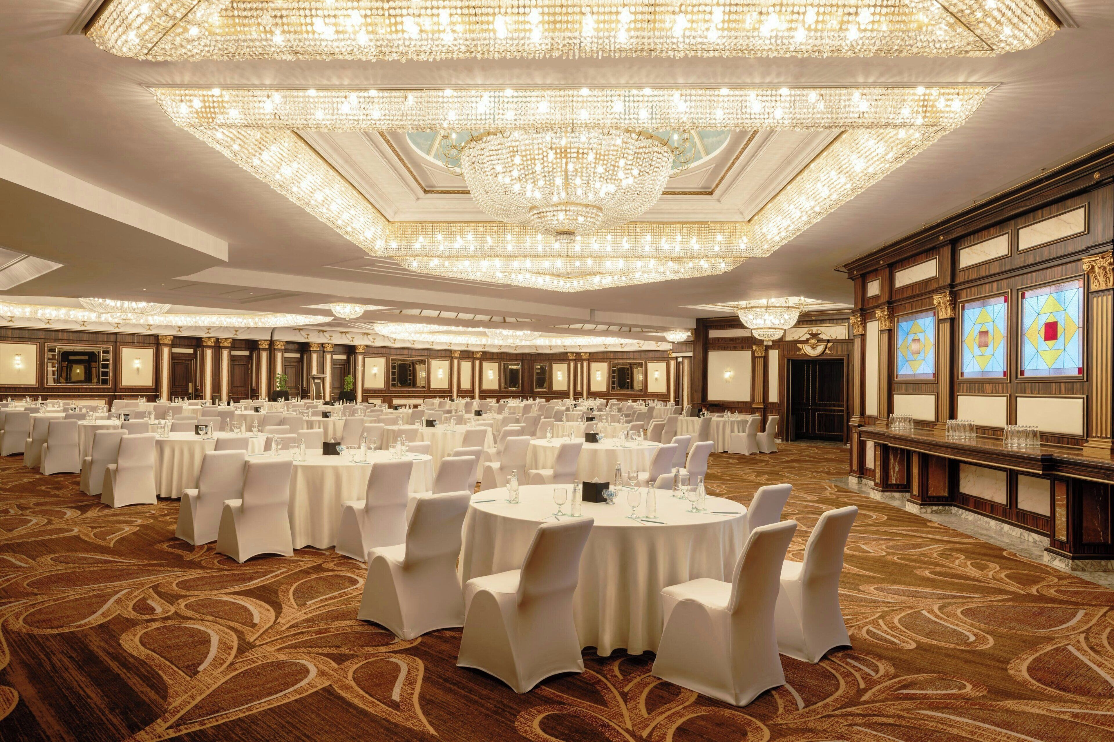 A banquet hall with round tables, white chairs, and chandeliers.