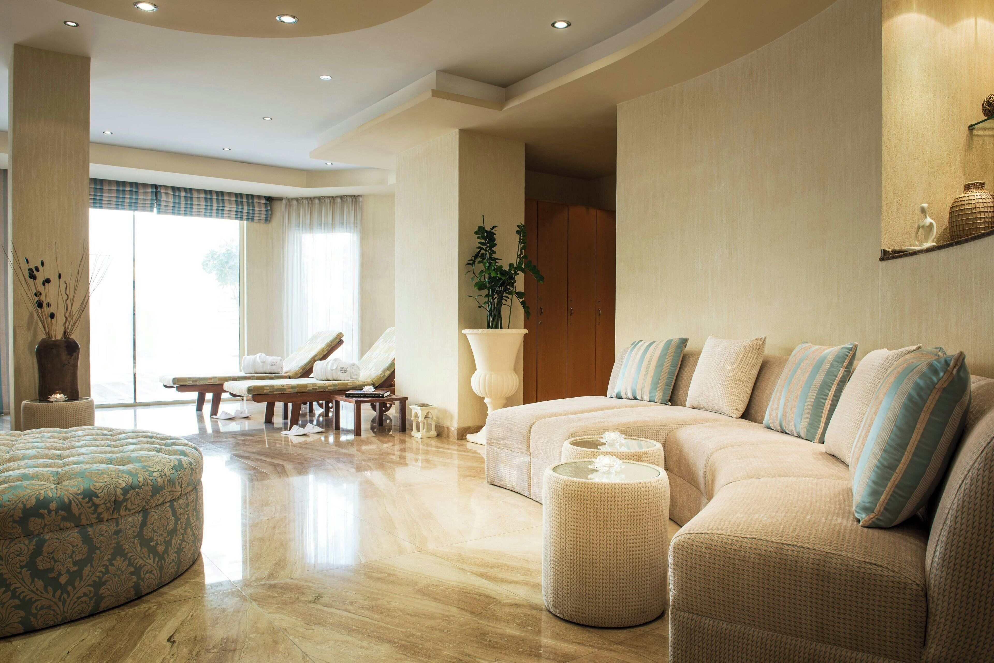 A spacious living area with a beige sofa, a patterned ottoman, and a large window with curtains.