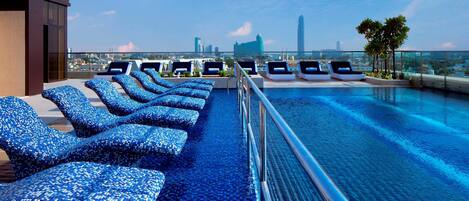 A rooftop infinity pool with blue mosaic lounge chairs and a city skyline view.