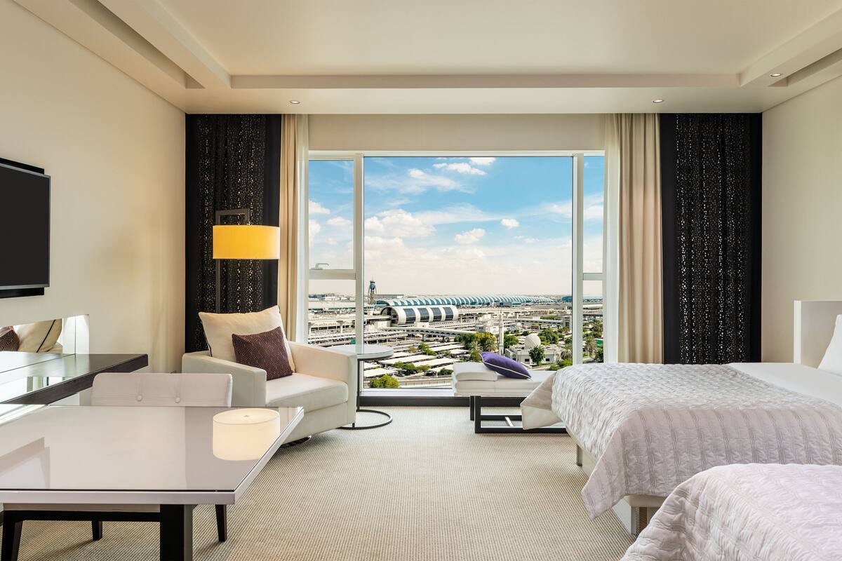A hotel room with a large window, a bed, a sofa, a table, and a TV.
