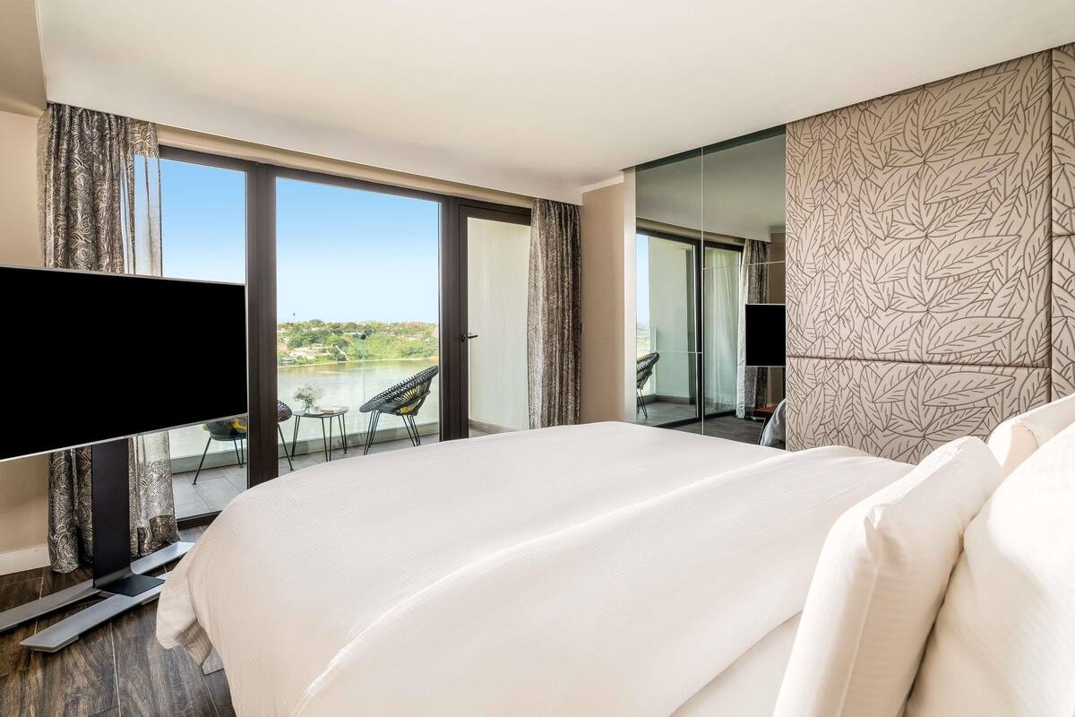 A hotel room with a large bed, a flat-screen TV, a balcony with a chair, and a view of a body of water.